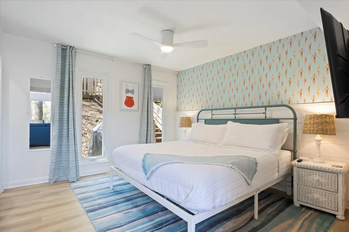 A cheerful bedroom featuring a bed with white bedding, a colorful accent wall, blue curtains, and a stylish rug. The space exudes comfort and freshness, ideal for unwinding after an active day.