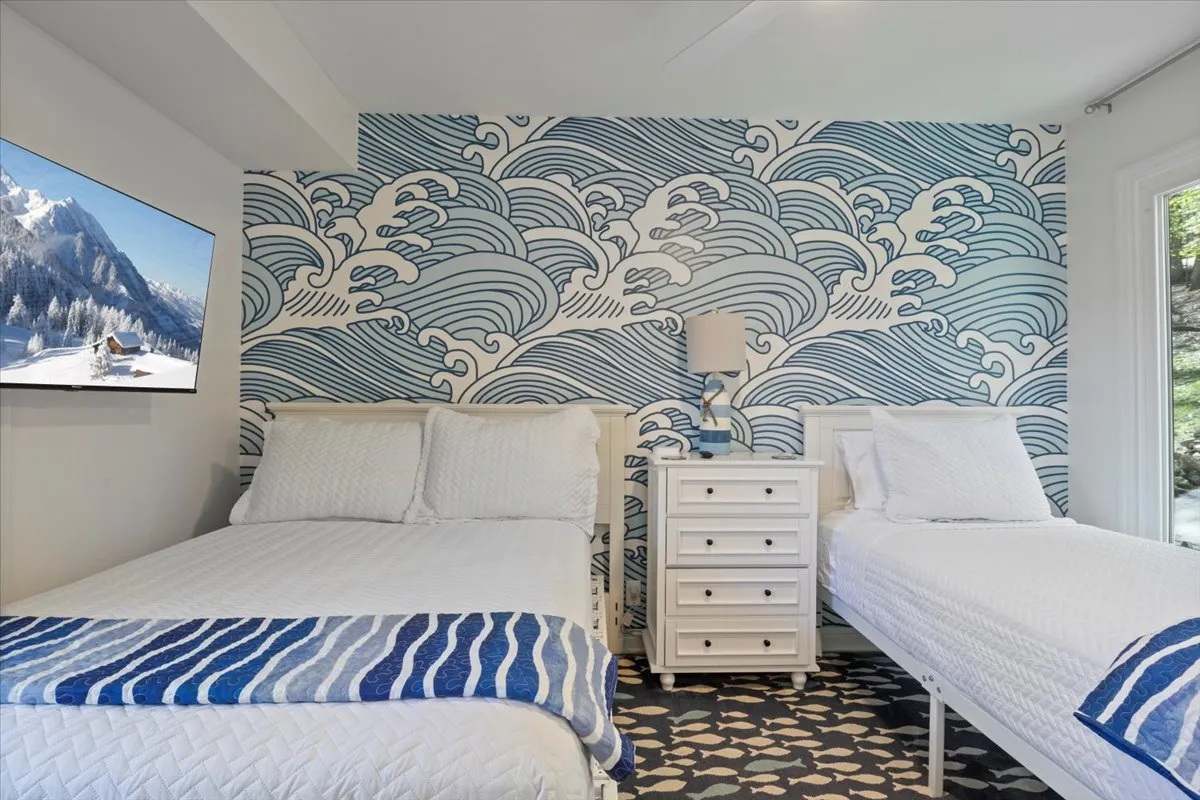 A tranquil bedroom with two beds dressed in white linens, a wave-patterned accent wall, blue accent blanket, and a nightstand with a lamp. A television is mounted on the wall, and large windows allow natural light to brighten the space, creating a peaceful, airy atmosphere.