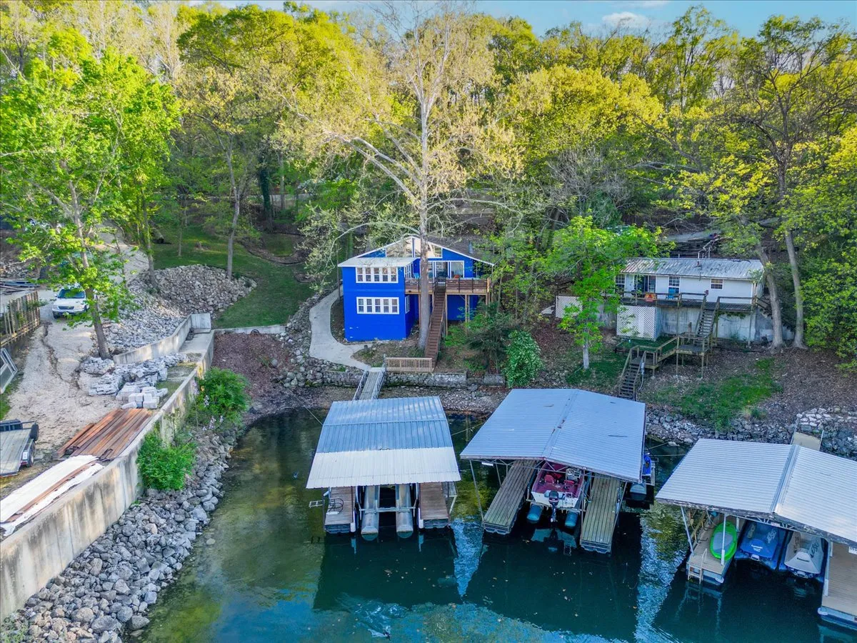 This image features the vibrant blue house with boat docks directly accessible from the property, surrounded by lush trees and calm lake waters. The setting clearly displays a private dock and slip, with the tranquil lake inviting boating and fishing activities, and the immediate proximity of water emphasizes the 'at your doorstep' promise.