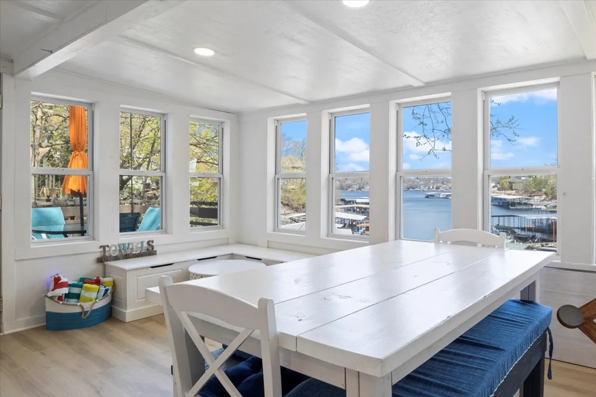 A bright dining area featuring a spacious table set beside large windows that frame an outdoor lake view. The space is filled with natural light and adorned with colorful towels, offering an inviting setting for family meals, sunset watching, and storytelling.