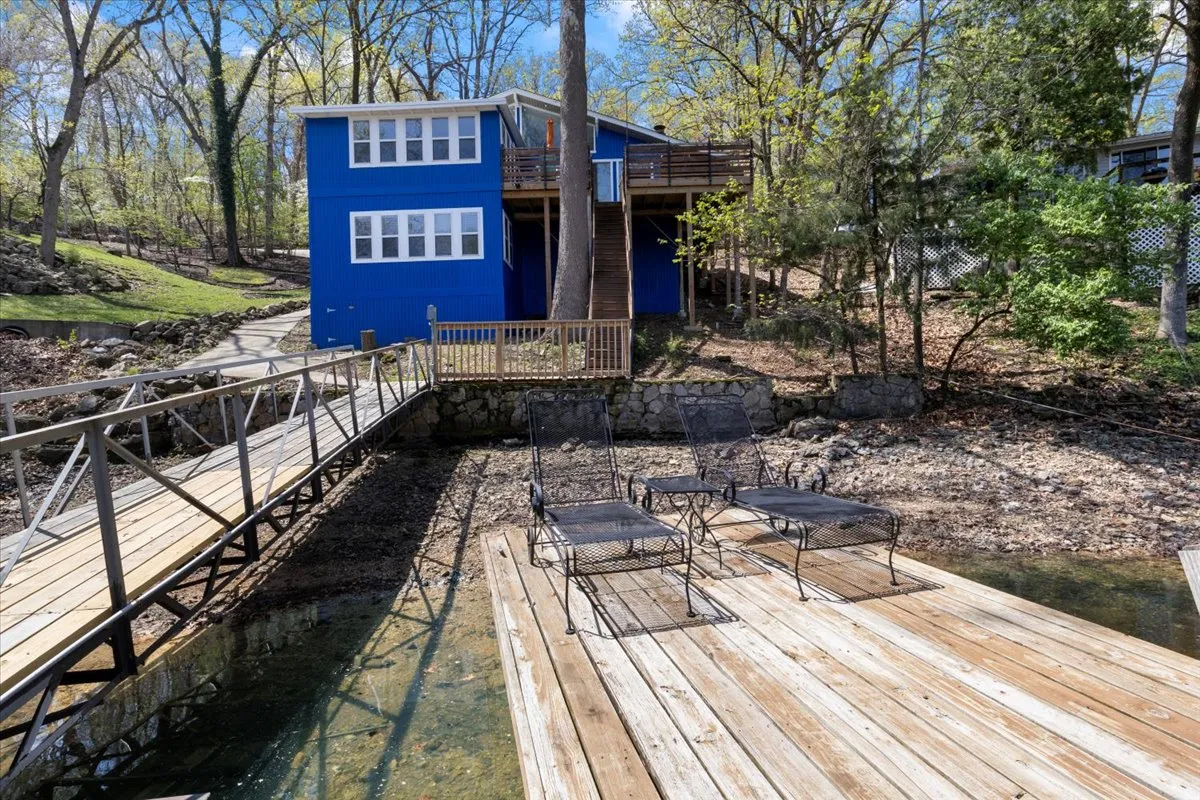 The bright blue house sits serenely by the water, featuring a spacious wooden deck with metal chairs and surrounded by lush, natural greenery. The home’s cheerful façade and peaceful setting evoke the perfect starting point for family gatherings and lakeside adventures.