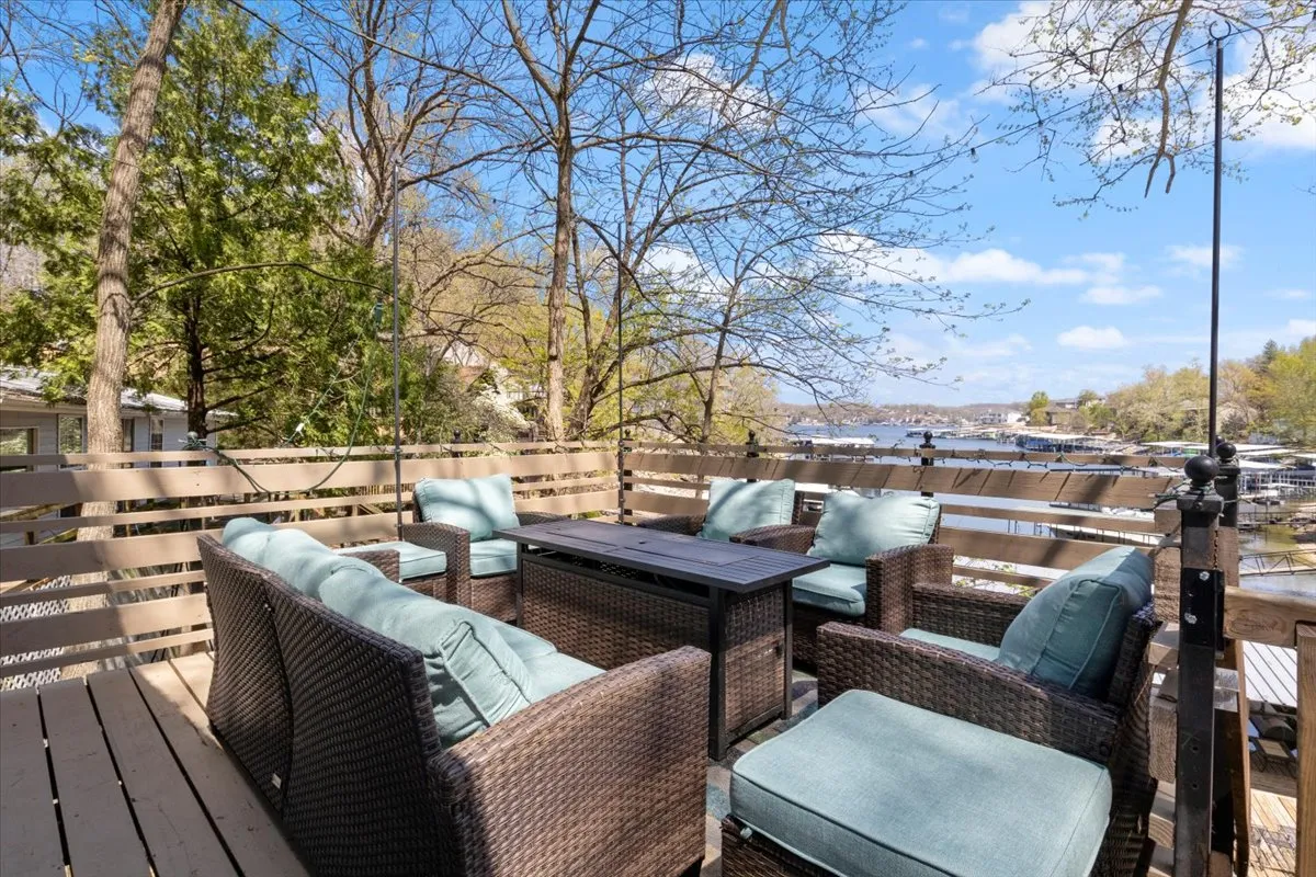 The image showcases a cozy outdoor seating area nestled beneath mature trees, overlooking serene waters of Lake of the Ozarks. The space invites relaxation with comfortable chairs arranged for conversation, and the tranquil lake view framed by lush foliage hints at both sunrise coffees and sunset gatherings.