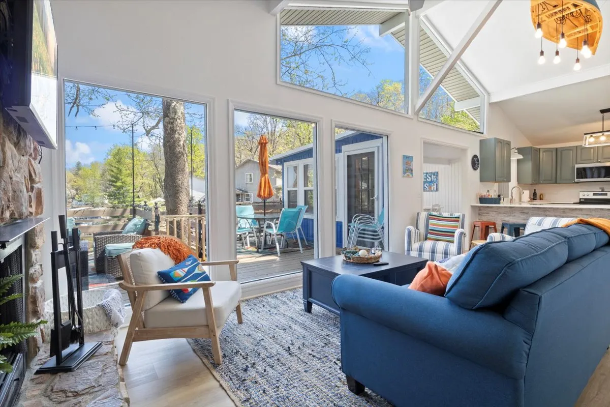 A sun-filled living room with large windows offering lake views, a comfortable blue sofa accented by decorative throw pillows, a central coffee table ideal for board games or refreshments, and a welcoming stone fireplace that anchors the space. The atmosphere is warm and inviting, perfect for gathering friends and family.