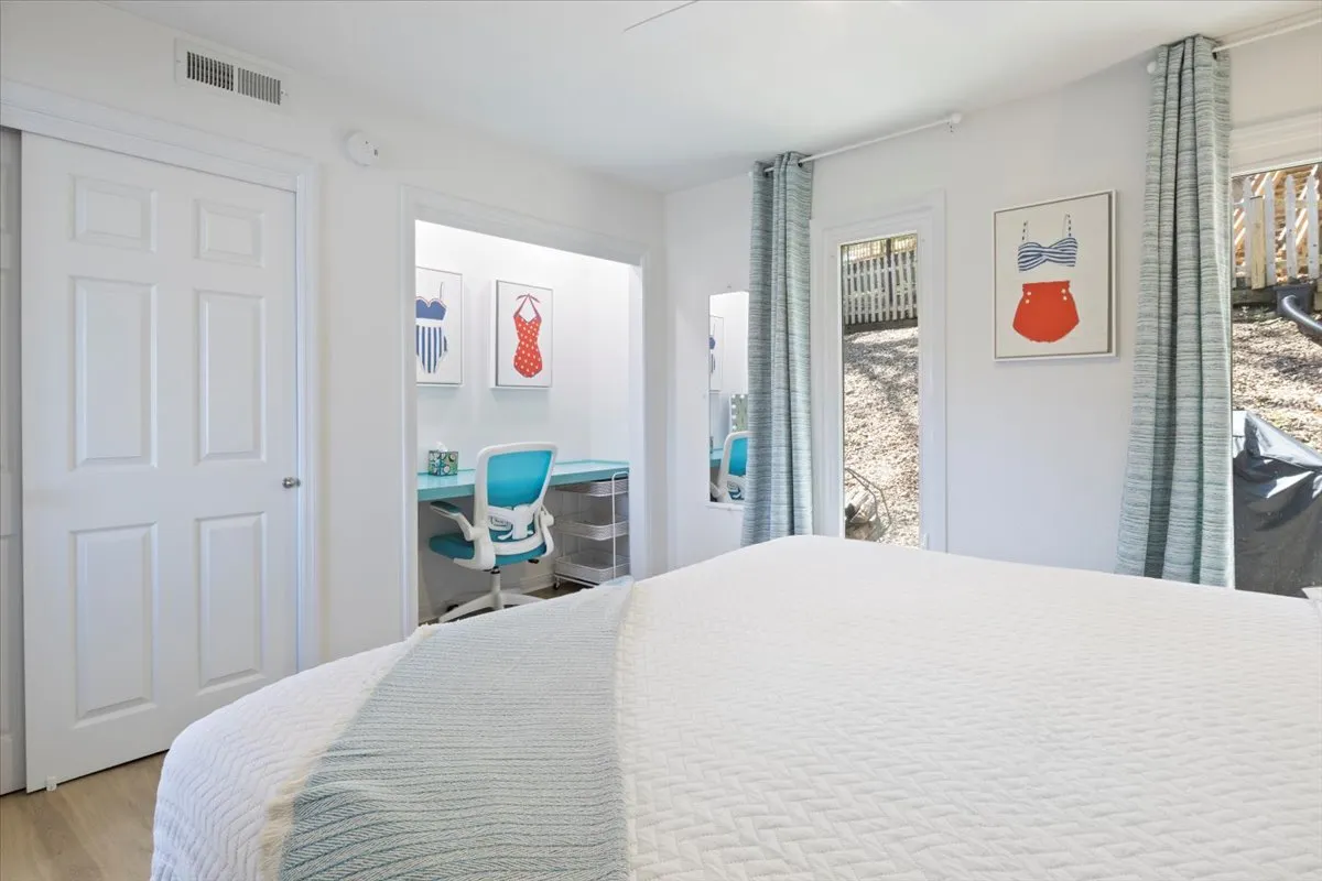 This image showcases a bedroom featuring a light blue desk and chair setup positioned by a window, with swimwear-themed artwork on the wall and a bed with a textured white coverlet. Sunlight fills the space, giving it a refreshing and playful atmosphere. The desk area is functional with a view outside, ideal for remote work or creative tasks.