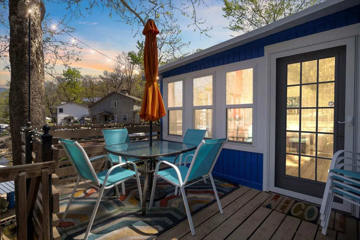 This image showcases a cheerful outdoor dining set complete with an orange umbrella, situated on a deck attached to a blue and white house. Decorative touches such as a patterned rug create a warm and inviting ambiance. The space is clearly designed for social gatherings and al fresco meals.