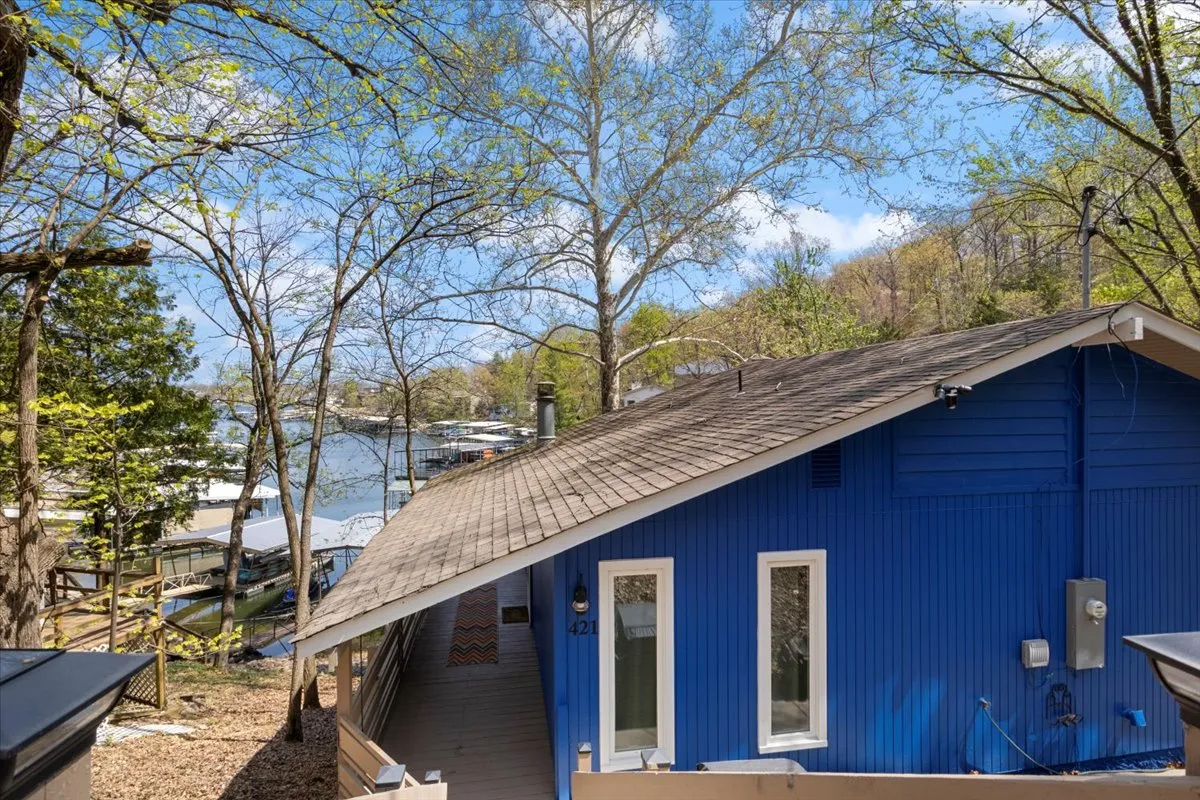 A wide exterior perspective featuring the bright blue house surrounded by trees, with a clear view of the lake and dock in the background. Large windows invite light into the living spaces, and the deck offers a space for morning coffee or evening gatherings with scenic water views.